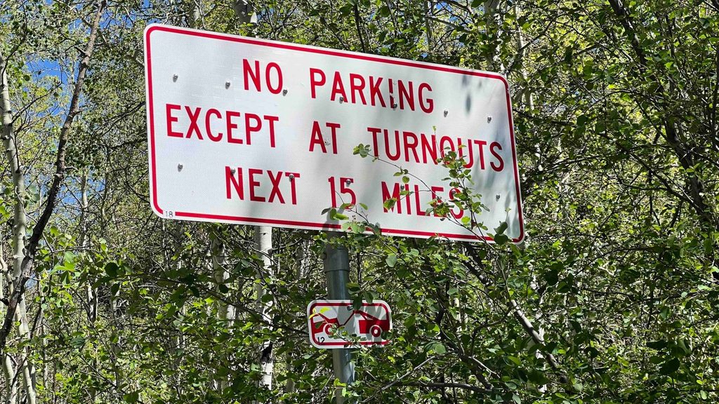 A sign surrounded by aspen trees reads, “No parking except at turnouts next 15 miles” with an illustration of a tow truck